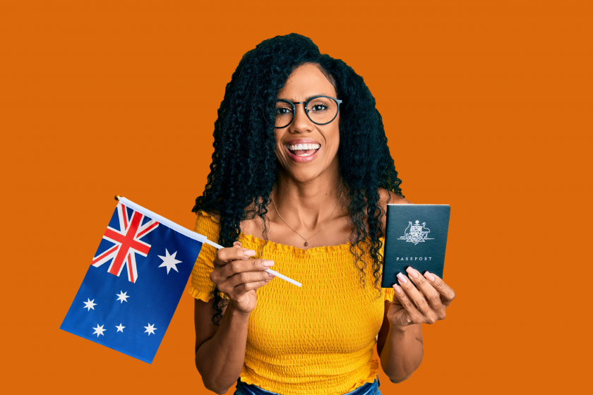 A young African Woman, dressed in a yellow shirt, jeans, and glasses is holding her passport and the Australian flag, smiling in front of an orange background.